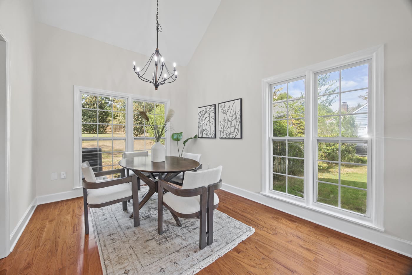 The dining area with a round table beneath an iron chandelier and two windows to the side yard.